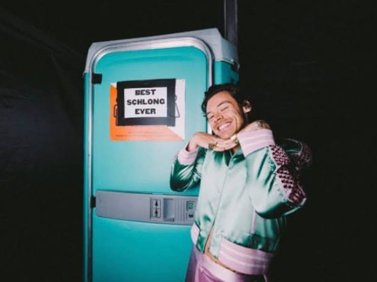 The former One Direction member posing near the restroom with the sign reading 'Best Schlong Ever'